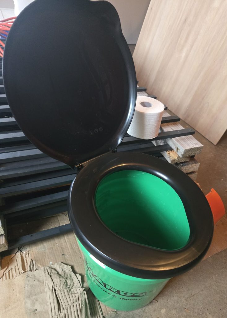 a green menards bucket with a black toilet lid on top and a roll of toilet paper next to it.